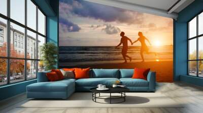 Happy couple running at sunset sea beach with holding hands. Honeymoon travel for spouses to tropical paradise country. Man and woman in love have fun in evening sunshine. Wall mural