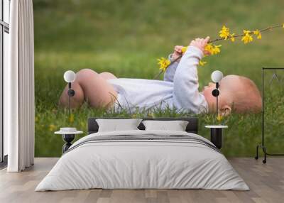 Little baby boy is lying on green gras and playing with yellow flower Wall mural