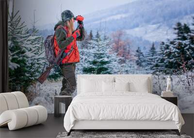 A young beautiful woman looks into the binocular with rifle on the shoulder ready for the hunt. There are snowy trees all around. Wall mural