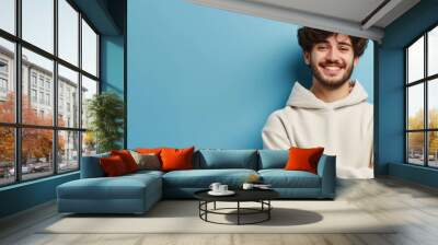 Portrait of happy man with curly hair smile toothily keeps arms down wears casual hoodie looks cheerful isolated over blue background being in good mood with copy space. Generative ai Wall mural