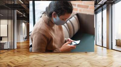 Mujer joven morena con jersey de cuello alto marrón con mascarilla azul sentada en un sofá verde mirando el teléfono móvil Wall mural