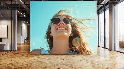 Portrait of a young Caucasian girl, wearing sunglasses, smiling against the sunny blue sky of a summer day. Wall mural