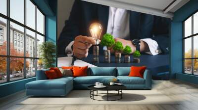 Businessman holding a light bulb and stacks of money growing with green trees. Concept of
pass and increase of renewable energy. Alternative sources of energy. Green energy, eco energy.  Wall mural