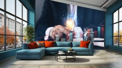 Businessman holding illuminated light bulb futuristic and stack of coins with gidital  graph. Business innovation technology. Wall mural