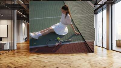 young woman sits on a tennis court, smiling and holding a tennis racket. The sun shines brightly, and the court is green Wall mural