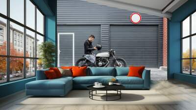 Handsome rider man take off black biker helmet while sit on classic style cafe racer motorcycle near industrial gates. Bike custom made in vintage garage. Brutal fun urban lifestyle. Outdoor portrait. Wall mural