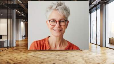 Middle-aged Caucasian woman with short gray hair and glasses, wearing an orange top, smiling, and looking at the camera against a plain light gray background Wall mural