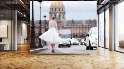 A beautiful young woman walks in a luxurious short white lace dress, high-heeled shoes, next to the road, in Paris, next to the Les Invalides Wall mural