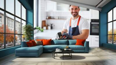 Smiling professional plumber in overalls with toolbox in modern white kitchen setting Wall mural