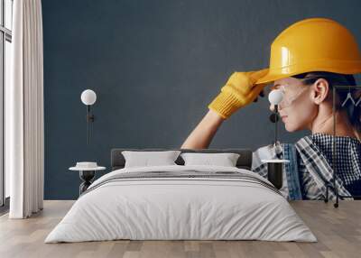 a strong-willed woman in a construction helmet, mittens, goggles and overalls is engaged in repair and construction work at home. concept of a strong and independent woman Wall mural