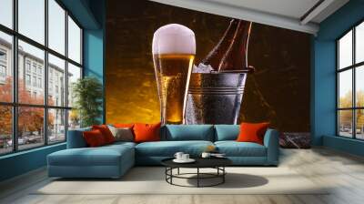 two bottles of beer in a bucket with ice and a glass of beer with lush foam next to a dark background Wall mural