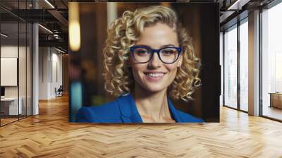 A woman with curly blond hair smiles while wearing blue eyeglasses Wall mural