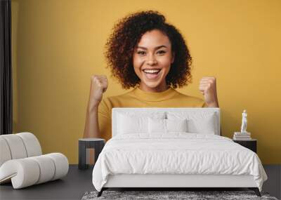 A woman with curly hair raises her fists in the air and smiles brightly against a yellow background Wall mural