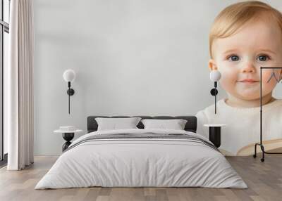  A baby gazes at the camera with a surprised expression, holding a book in front of a pristine white backdrop Wall mural