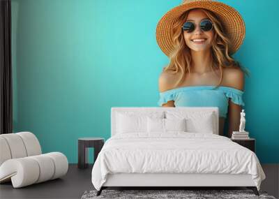 A cheerful woman in summer attire, wearing a straw hat and sunglasses, stands against a bright turquoise wall, exuding joy and carefree vibes Wall mural