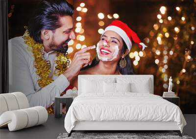 Happy Indian man applying cake cream on woman's face on Christmas night Wall mural