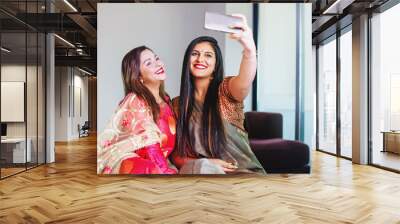 Two beautiful Indian women in ethnic clothes taking selfie on mobile phone at home Wall mural