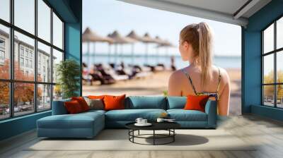 Young woman with ponytail, blonde hair is looking on the sea horizon, vacation mood, summer travel concept. Beach umbrellas on the background Wall mural