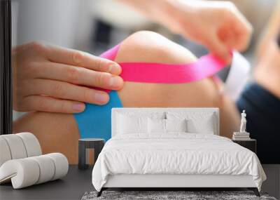 Kinesiology taping treatment with blue and pink tape on athlete patient injured knee. Woman hands apply kinesio treatment after sports muscle injury. Wall mural