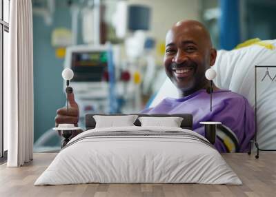 Smiling male patient showing thumbs up. Happy middle aged black bald man after chemotherapy in hospital bed. African american patient in purple gown. Cancer treatment success concept. Wall mural