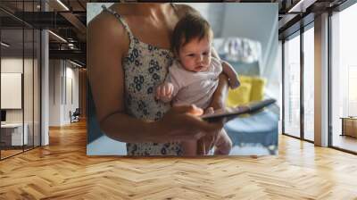 Adult woman mother holding three months old baby while using looking mobile phone for internet browsing or social network texting in room at home domestic life motherhood and parenting busy concept Wall mural