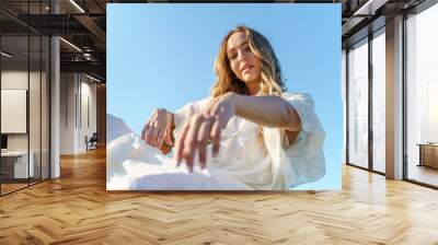 Low angle view of adult caucasian woman female model in white dress looking to the camera while standing or sitting against a blue sky in sunny summer day Wall mural