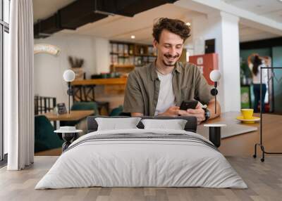 one caucasian man young adult millennial male using mobile phone sit at table in cafe at the table alone happy smile confident having online chat using internet for text messages or social network Wall mural