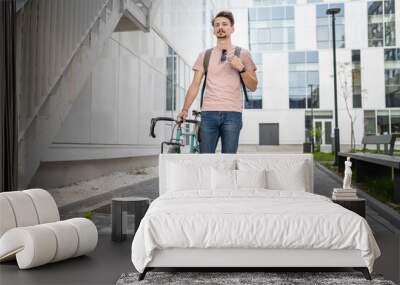 One man young adult male with brown hair and mustaches walking by the building with rucksack on his back and bicycle happy smile joyful real people copy space full length front view Wall mural