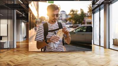 One man young adult modern caucasian male in the city in sunny day walk and eat sandwich fast food concept urban life copy space tourist eating on the street real person Wall mural