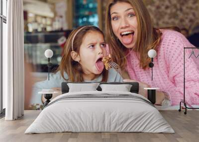 Portrait of small caucasian girl with mouth opened sitting by the table at restaurant while her mother is feeding brunette daughter holding fork and food in day posing looking to the camera Wall mural