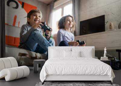 Two children small caucasian brother and sister happy children siblings boy and girl playing video game console using joystick or controller while sitting at home real people family leisure concept Wall mural