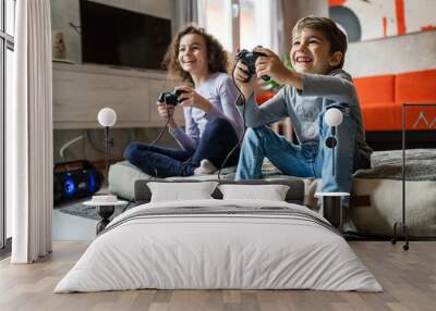 Two children small caucasian brother and sister happy children siblings boy and girl playing video game console using joystick or controller while sitting at home real people family leisure concept Wall mural