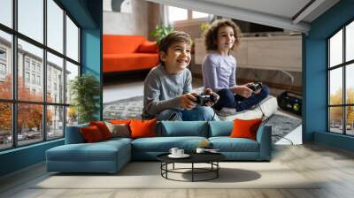 Two children small caucasian brother and sister happy children siblings boy and girl playing video game console using joystick or controller while sitting at home real people family leisure concept Wall mural