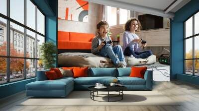 Two children small caucasian brother and sister happy children siblings boy and girl playing video game console using joystick or controller while sitting at home real people family leisure concept Wall mural