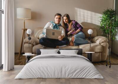 Two people man and woman young adult caucasian couple boyfriend and girlfriend or married husband and wife sitting on the sofa bed at home using mobile phone and laptop for work or internet browsing Wall mural