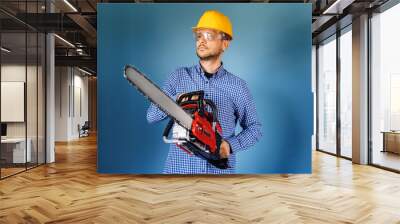 Waist up of adult caucasian man wearing protective helmet and glasses standing in front of the blue wall hold chainsaw tool selective focus on chainsaw Wall mural