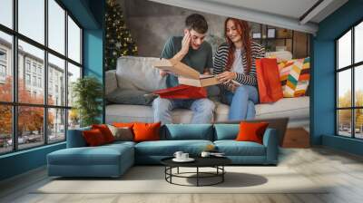 Young cheerful couple open gift from shopping bags during video call Wall mural