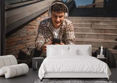 Young happy man sit on the stairs with headphones and listen music Wall mural