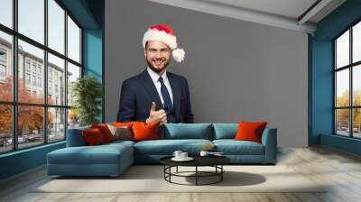 Portrait of cute business guy successful businessman wearing suit and Christmas hat showing thumb up against grey studio wall background. Wall mural