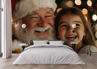 A joyful Santa Claus, wearing his classic red suit and white beard, shares a hearty laugh with a gleeful young girl by a sparkling Christmas tree, evoking holiday cheer. Wall mural