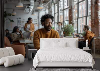 A man in a brown sweater works on his laptop in a bright, modern open office space with other people collaborating in the background, creating a vibrant work environment. Wall mural