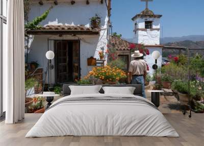 A man wearing casual attire stands in front of a rustic church adorned with vibrant potted plants and flowers, set against a backdrop of picturesque hills and traditional houses. Wall mural