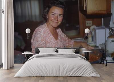 A smiling woman stands in a cozy kitchen stirring a pan of tomato sauce, with light wood cabinets and a welcoming atmosphere, highlighting culinary passion and warmth. Wall mural