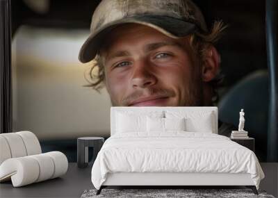 A young man with a rugged look, wearing a cap inside his vehicle, smiles contentedly at the camera, conveying a sense of ease and rugged outdoorsy charm. Wall mural