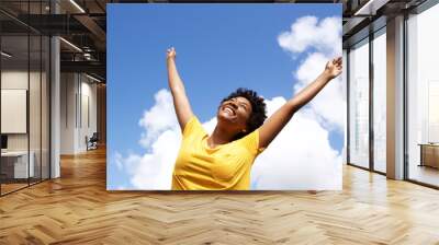 cheerful young woman with hands raised towards sky Wall mural