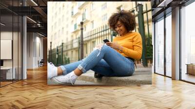 cool young african american woman sitting outside on street with mobile phone Wall mural