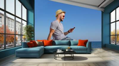 Happy young man sitting by the sea with mobile phone Wall mural