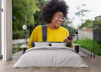 smiling young black woman with glasses and bag looking at cellphone outdoors Wall mural