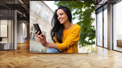 smiling young North African beautiful woman sitting outside in city looking at cellphone Wall mural