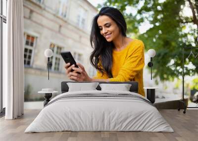 smiling young North African beautiful woman sitting outside in city looking at mobile phone Wall mural